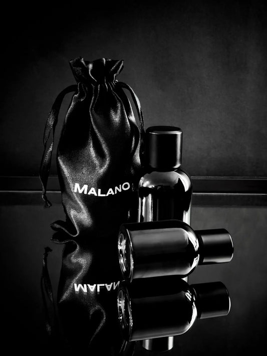 two perfume bottles sitting reflected on a mirror, one perfume bottle sitting up straight and the other on it's side. There is a black Malano perfume bag sitting behind them and all are softly lit against a dark grey and black background. 