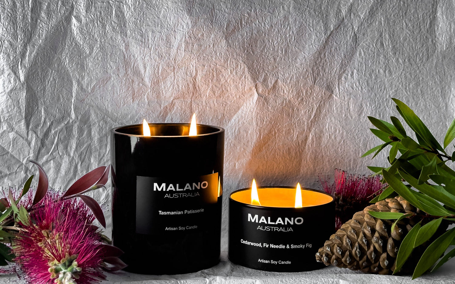 Two artisan soy wax candles hand poured by Malano sitting against a white textured background amongst some Australian flora including beautiful red bottle brush, eucalyptus and some pine cones. The scented candles; Tasmanian Patisserie which smells like a bakery and Cedarwood, Fir Needle and Smoky fig which smells like the wilderness, are lit and softly illuminating the scene they are nestled within. 
