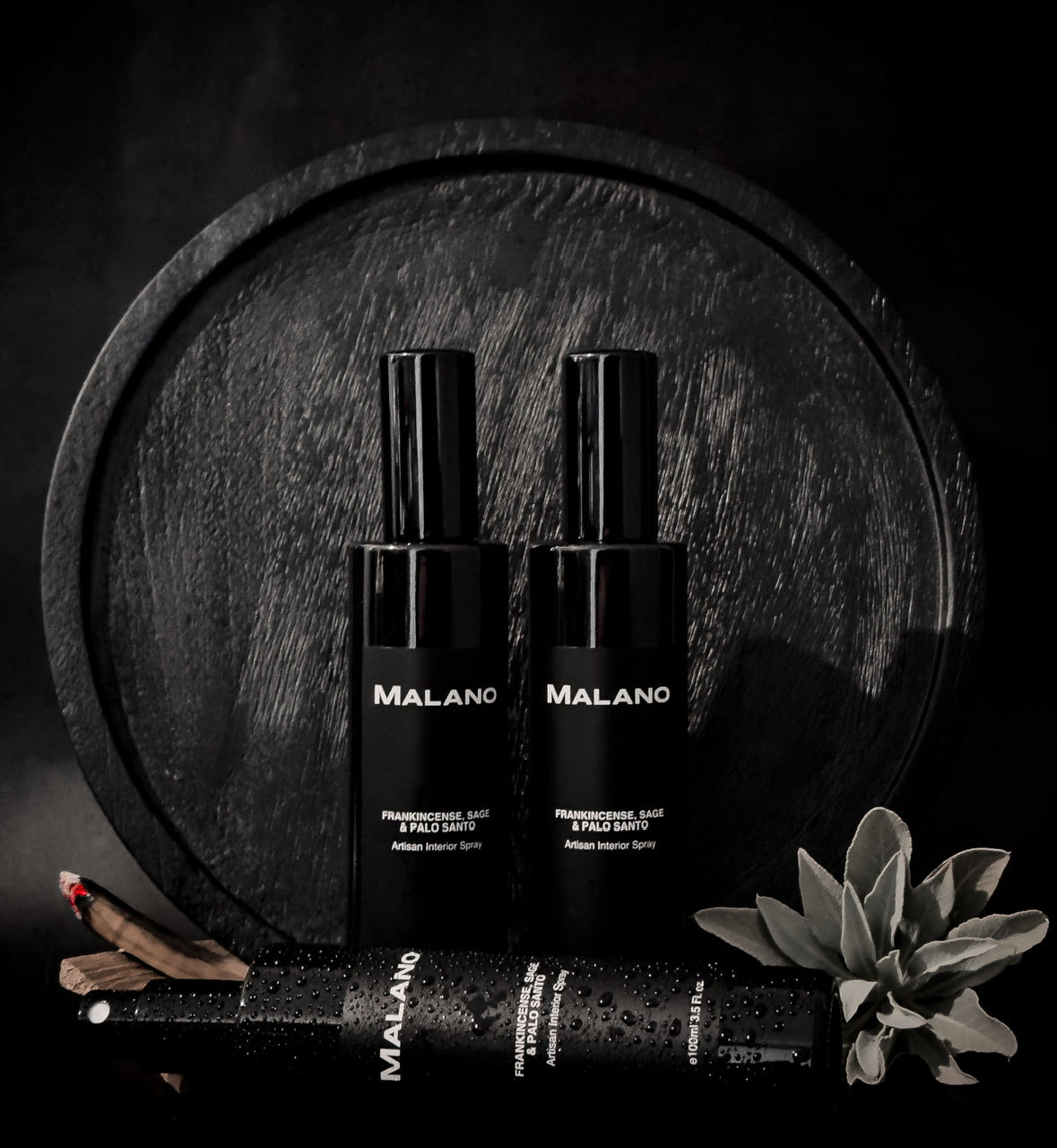 3 Spray Bottles of Frankincense, Sage and Palo Santo interior spray by Malano, two standing and one on its side covered in drops of water are sitting in front of a circular black textured wooden board and softly lit against a black background. To the right side is a sprig of sage and to the left side, three sticks of Palo Santo with one lit and smoke wafting upwards.
