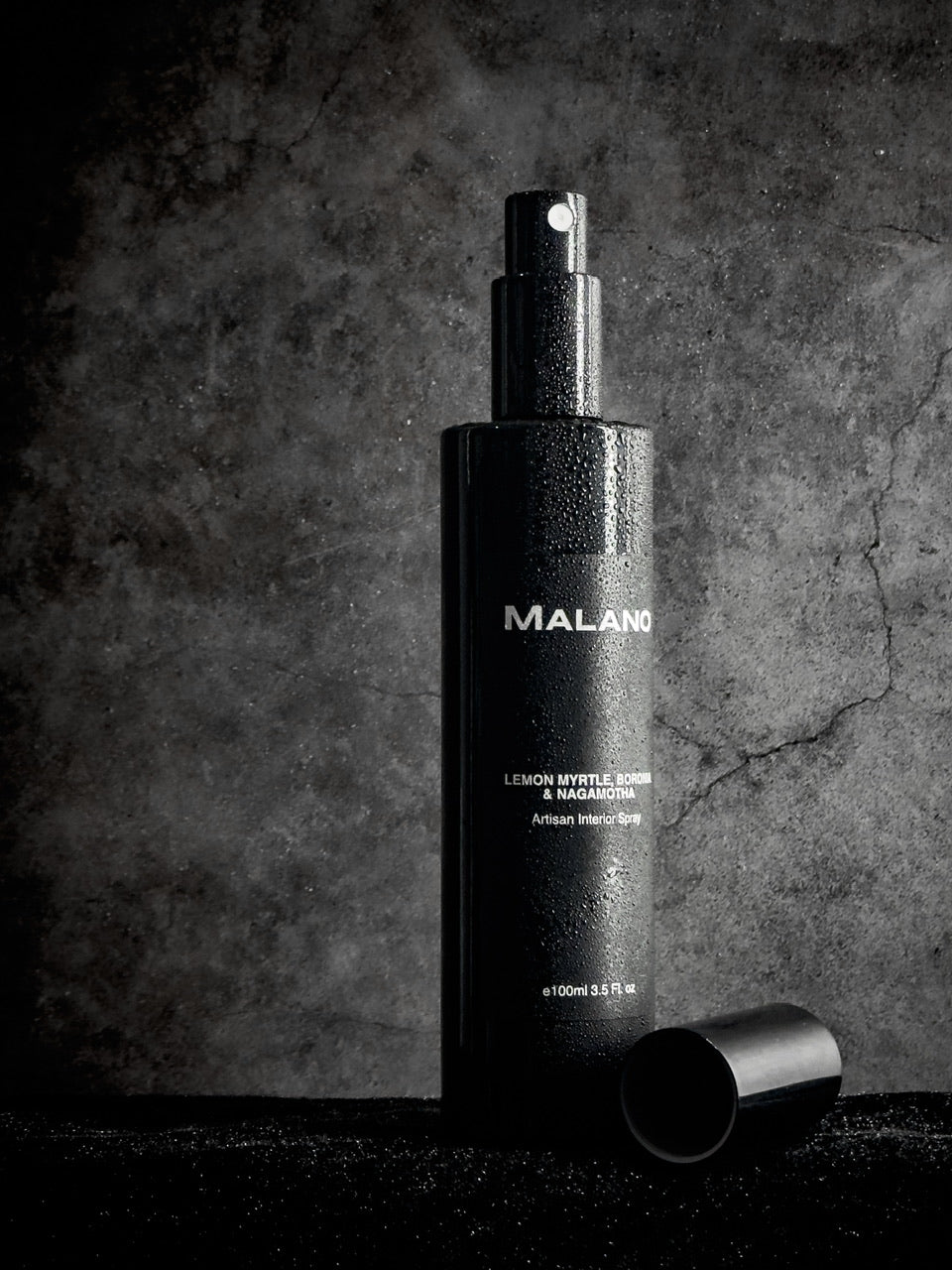 A tall, slim black bottle of Lemon Myrtle, Boronia and Nagamotha Interior Spray by Malano stands lightly lit against a grey concrete background, covered by a fine mist of liquid shimmering against the light. 