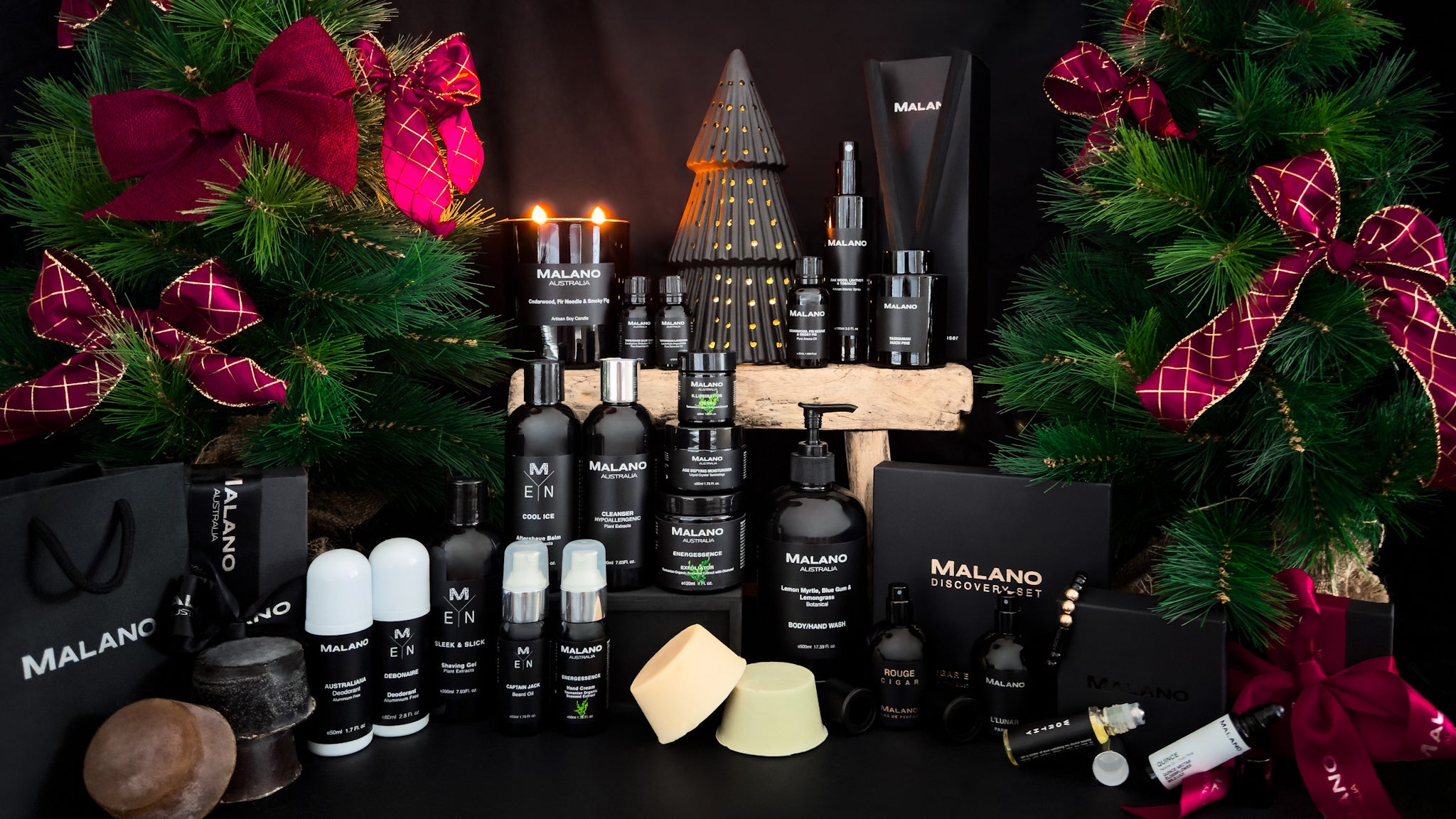 A Christmas image with 2 green pine trees decorated with burgundy and gold bows that site wither side of a showcase of Malano’s range of handcrafted, organic, plant-based products.