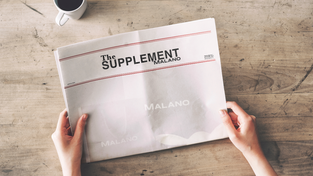 Person holding a newspaper titled 'The Supplement Malano' on a wooden table with a cup of coffee.