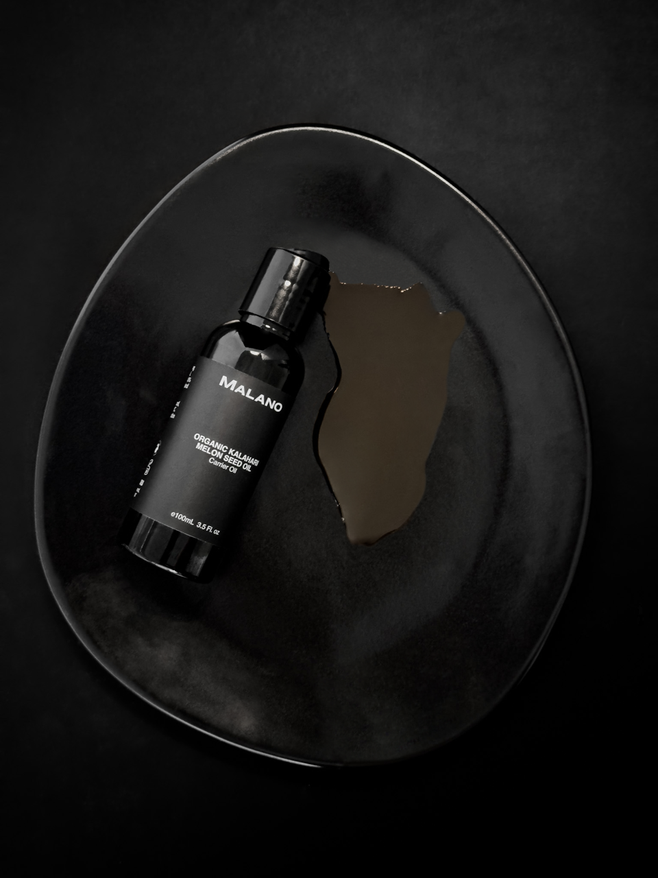 A Bottle of MALANO’s organic kalahari seed Oil in a black bottle with a capacity of 100mL. The oil is spilling out onto  a black egg shaped plate which is all laying against a dark background