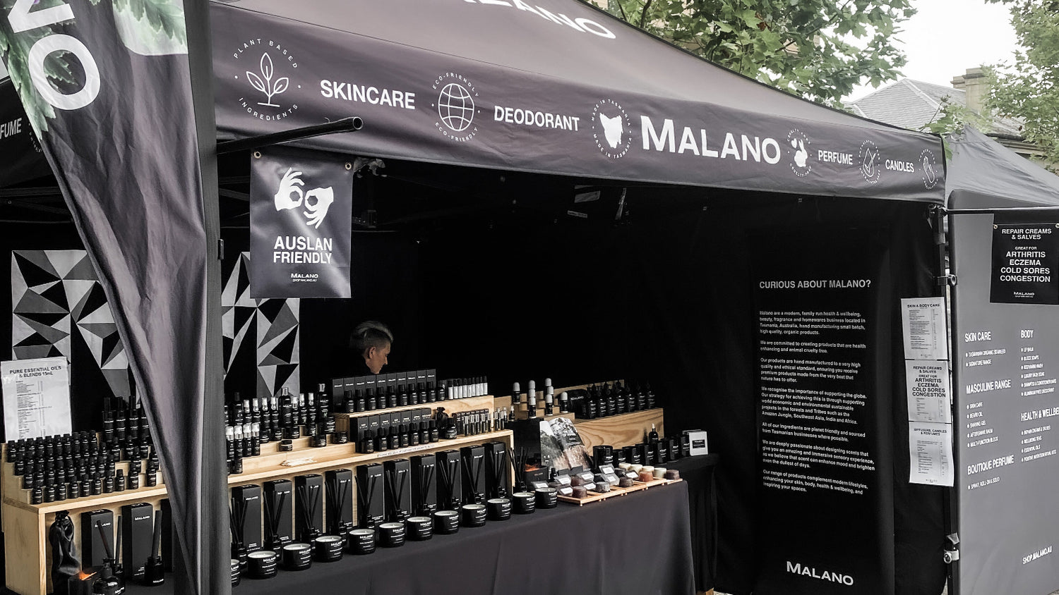 Malano skincare, body care, home fragrance,  and perfume products displayed under a black Malano tent with a pink floral banner, at Salamanca Market site 84-84 on Saturdays