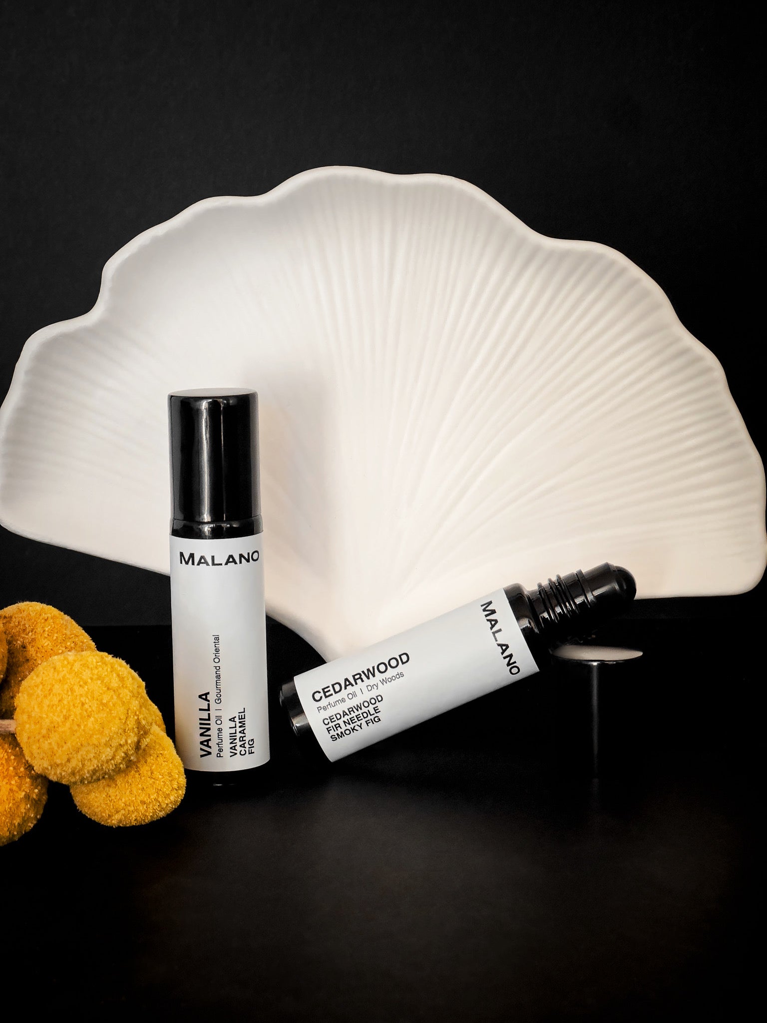 Two MALANO roll-on perfume oils—Vanilla and Cedarwood—elegantly displayed against a sculptural white fan backdrop. The Vanilla bottle stands upright, while Cedarwood lies open with the roller exposed. Soft yellow billy buttons add a natural touch on the side, all set against a sleek black background for a minimal, modern aesthetic.