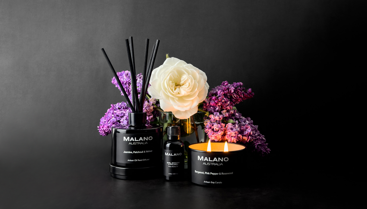 A collection of Malano scents of spring including a reed diffuser, pure aroma oil and candle. The collection has a white rose and 4 purple lilac flowers behind them and all are softly lit from the left hand side against a dark grey and black bacground.