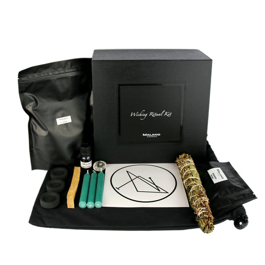 A Wishing Positive Intention Ritual Kit by Malano with it’s box and contents layed out in a tidy and organised manner including a bag of White Rock Salt, Anointing Oil, Charcoal, a stick of Palo Santo, three green candles, a gem stone, wishing sigil, Tree Fern, Tallowood, Tea Tree and Wattle smudge stick and Wishing Resin. These elements are sitting ontop of a black drawstring bag and against a white background.